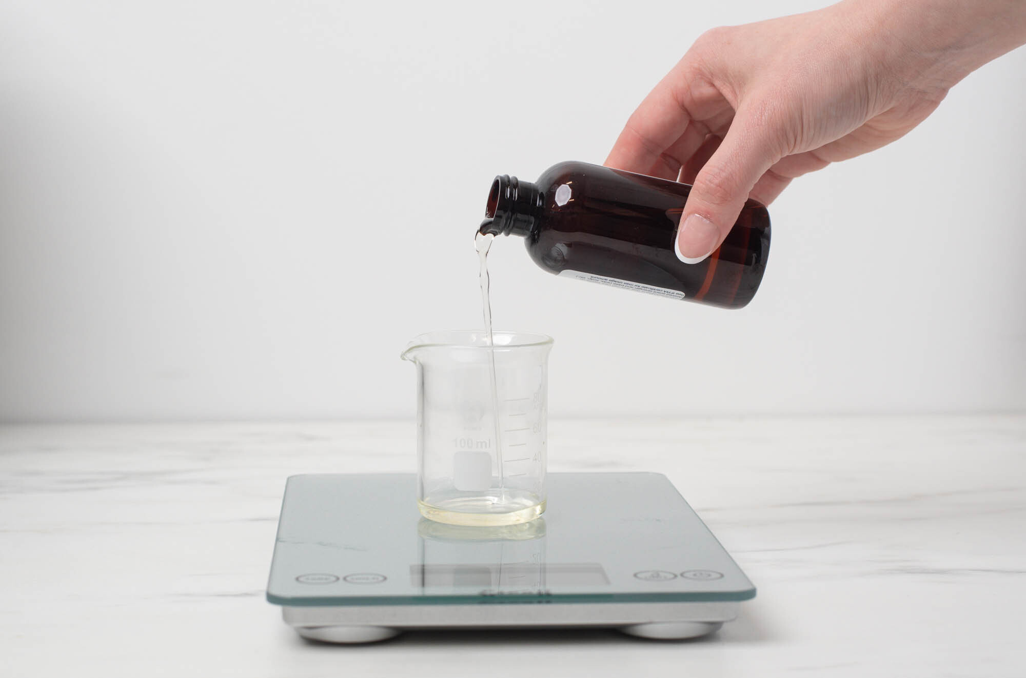 A hand pouring fragrance oil from a 4 ounce bottle into a glass measuring cup on a scale.